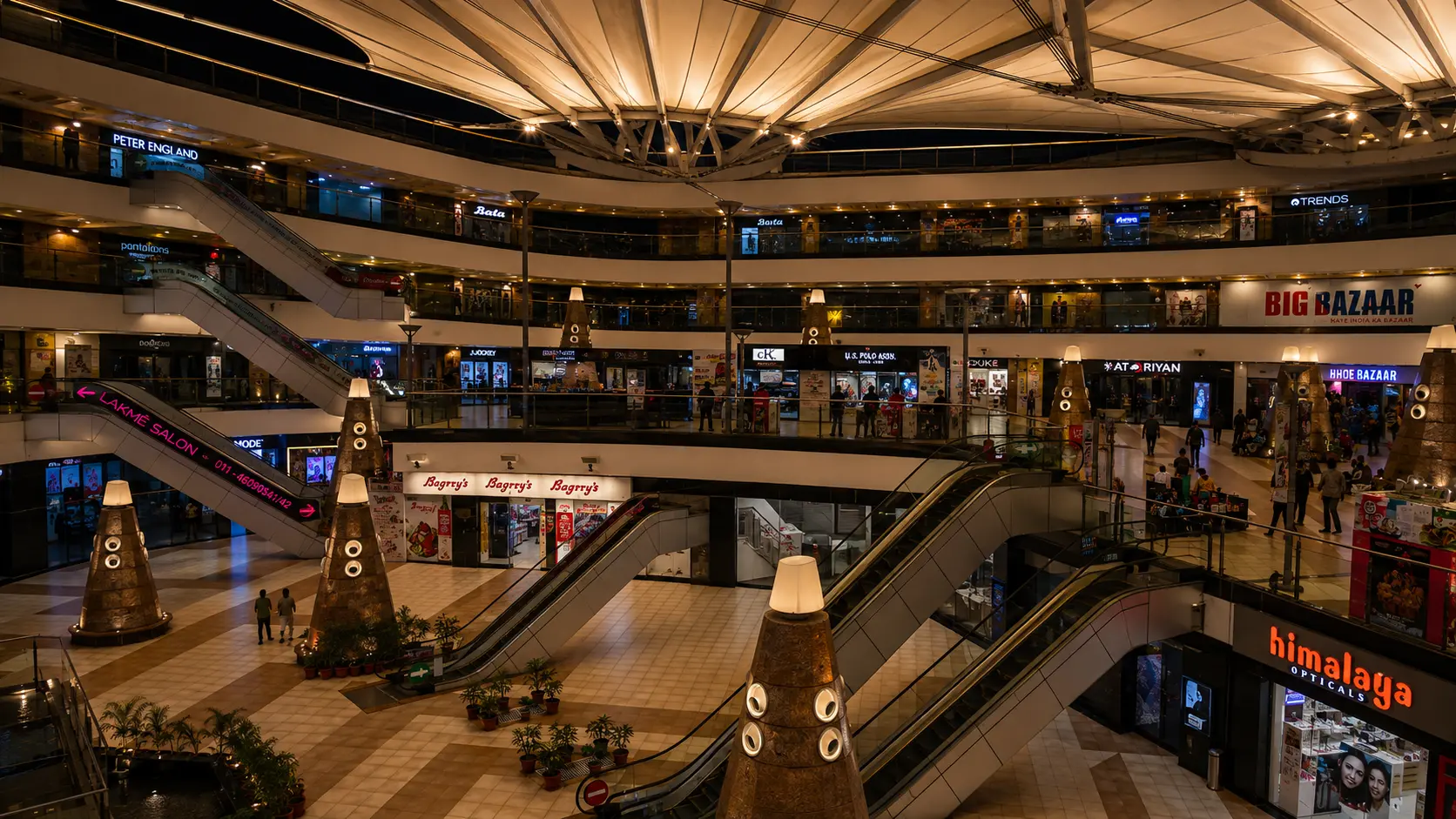 Vasant Square Mall Vasant Kunj tensile canopy at night, escalators and Bagrry's, Big Bazaar and Bata storefronts lit beneath it, Ekra Decor